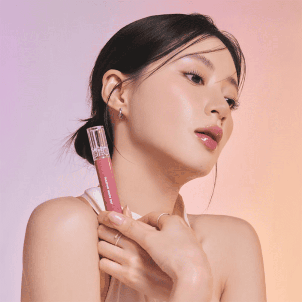 Udsnit af en kvinde med smuk hud, der holder en blush-penne i hånden, mod en pastel pink-orange baggrund. Perfekt til skønheds- og makeup-entusiaster, der ønsker glat og strålende hud.