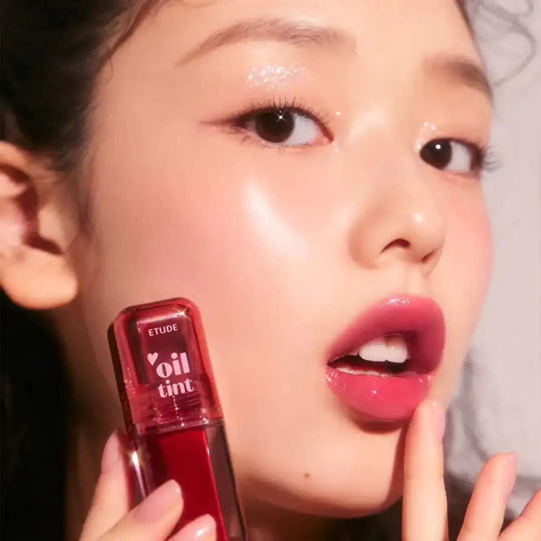Nærbillede af en ung kvinde med glansfulde læber og glittrende øjenbryn, der holder en rød ETUDE Oil Tint lip gloss. Perfekt til naturlige skønhedslooks, lip gloss og makeup inspiration.